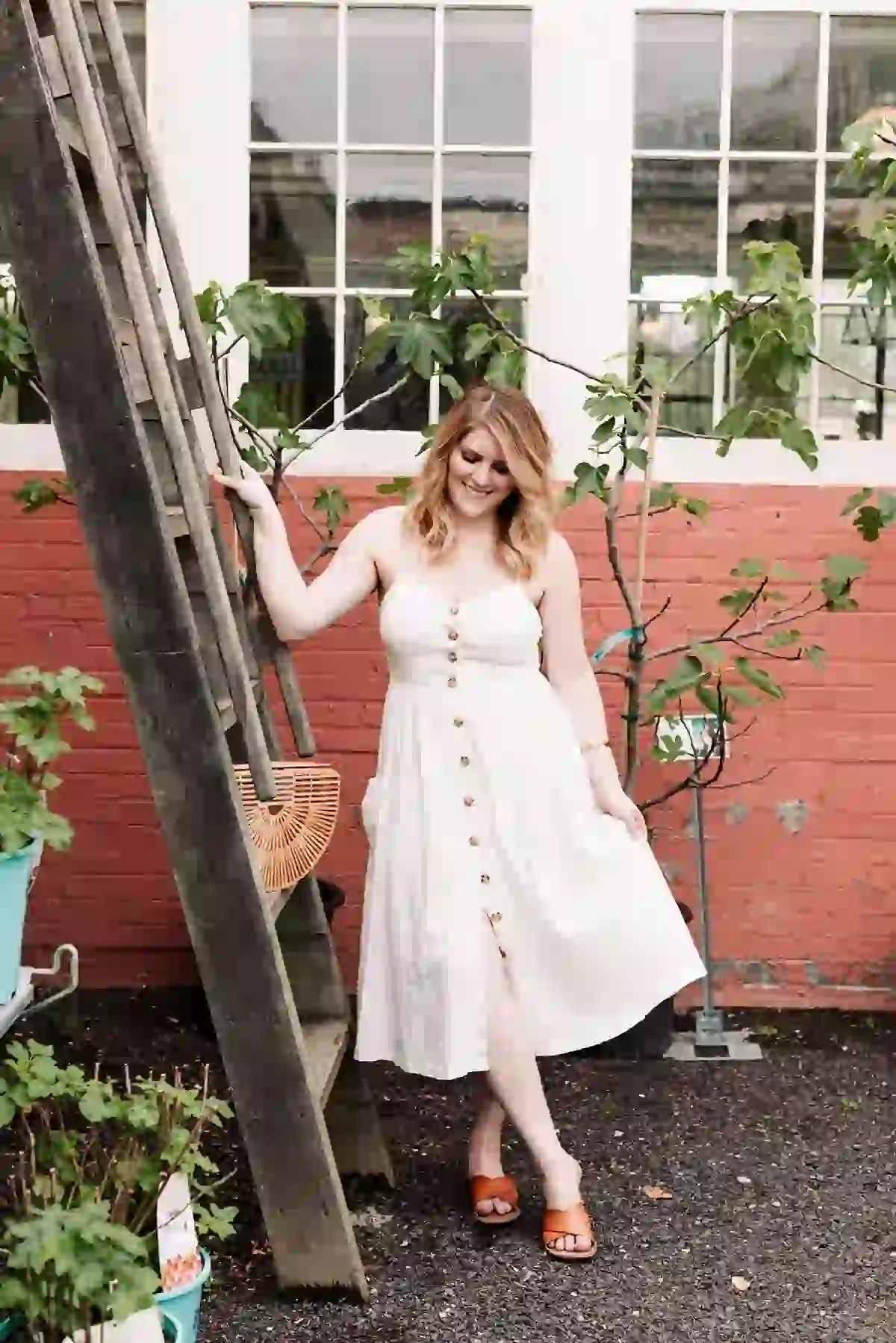 Woman in off-white day dress and brown sandals smiling as she stands outside in front of red bricks and light green foliage