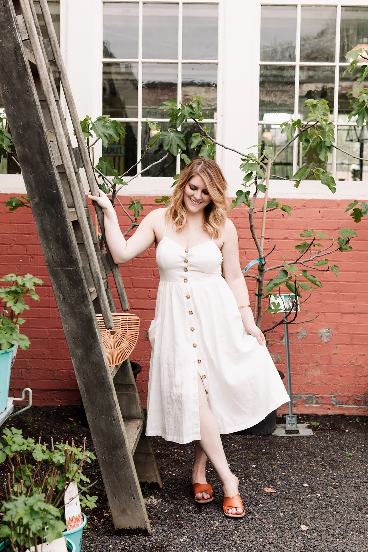 Woman in off-white day dress and brown sandals smiling as she stands outside in front of red bricks and light green foliage