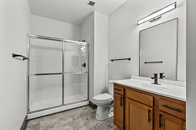 Renovated bathroom with glass shower door, gray tile floor, wood cabinetry, light countertop, and oversized vcanity mirror with even lighting